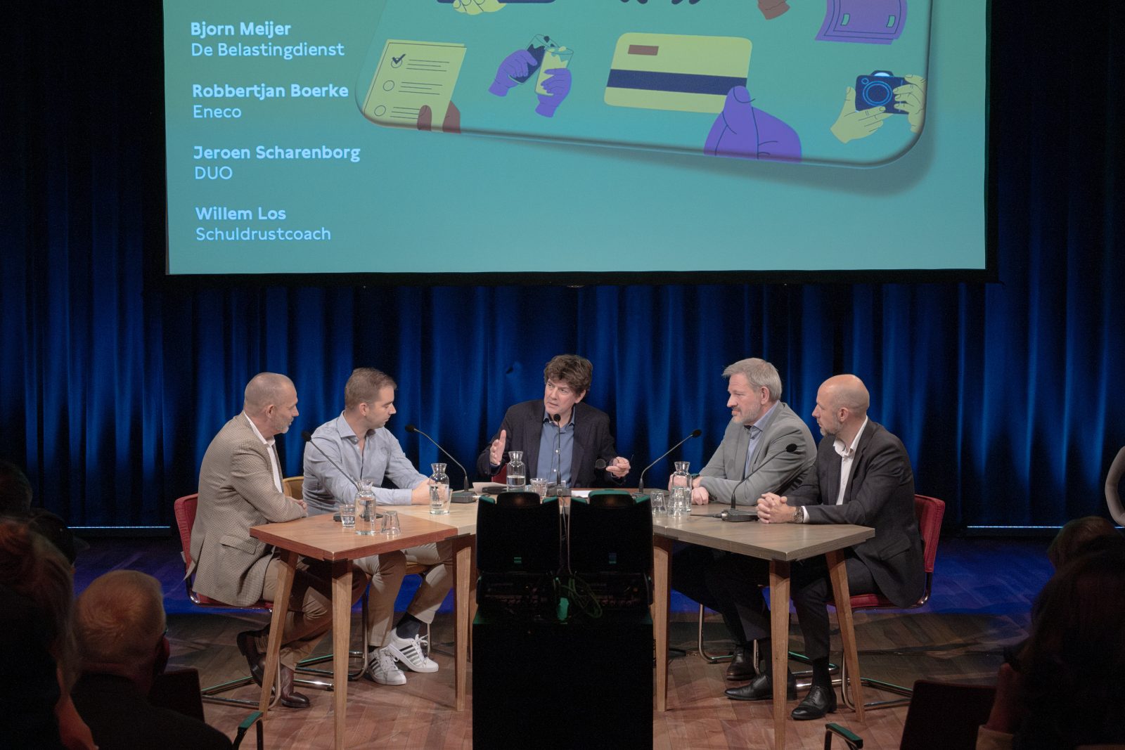 Foto van de bijeenkomst over schuldhulp aan jongeren. Aan de tafel zitten vijf personen. Op een groot scherm staat de namen van de deelnemers: Bjorn Meijer Belastingdienst, Robbertjan Boerke Eneco, Jeroen Scharenborg DUO, Willem Los Schuldrustcoach. Op de foto ook wat mensen uit het publiek.
