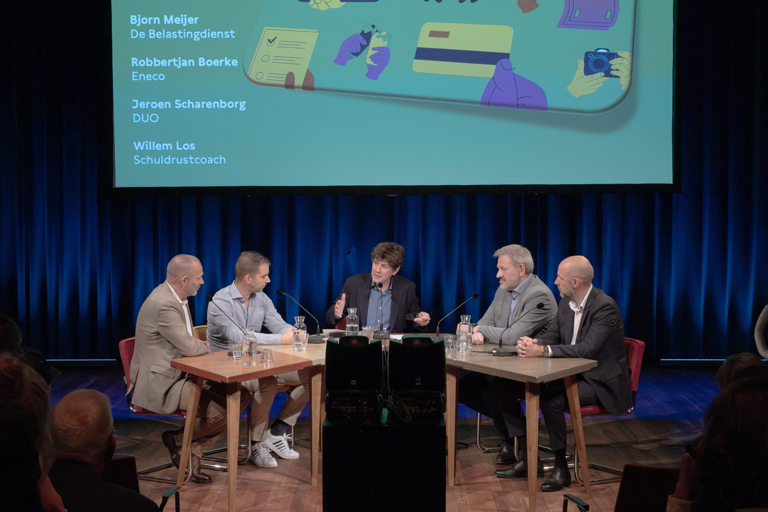 Foto van de bijeenkomst over schuldhulp aan jongeren. Aan de tafel zitten vijf personen. Op een groot scherm staat de namen van de deelnemers: Bjorn Meijer Belastingdienst, Robbertjan Boerke Eneco, Jeroen Scharenborg DUO, Willem Los Schuldrustcoach. Op de foto ook wat mensen uit het publiek.