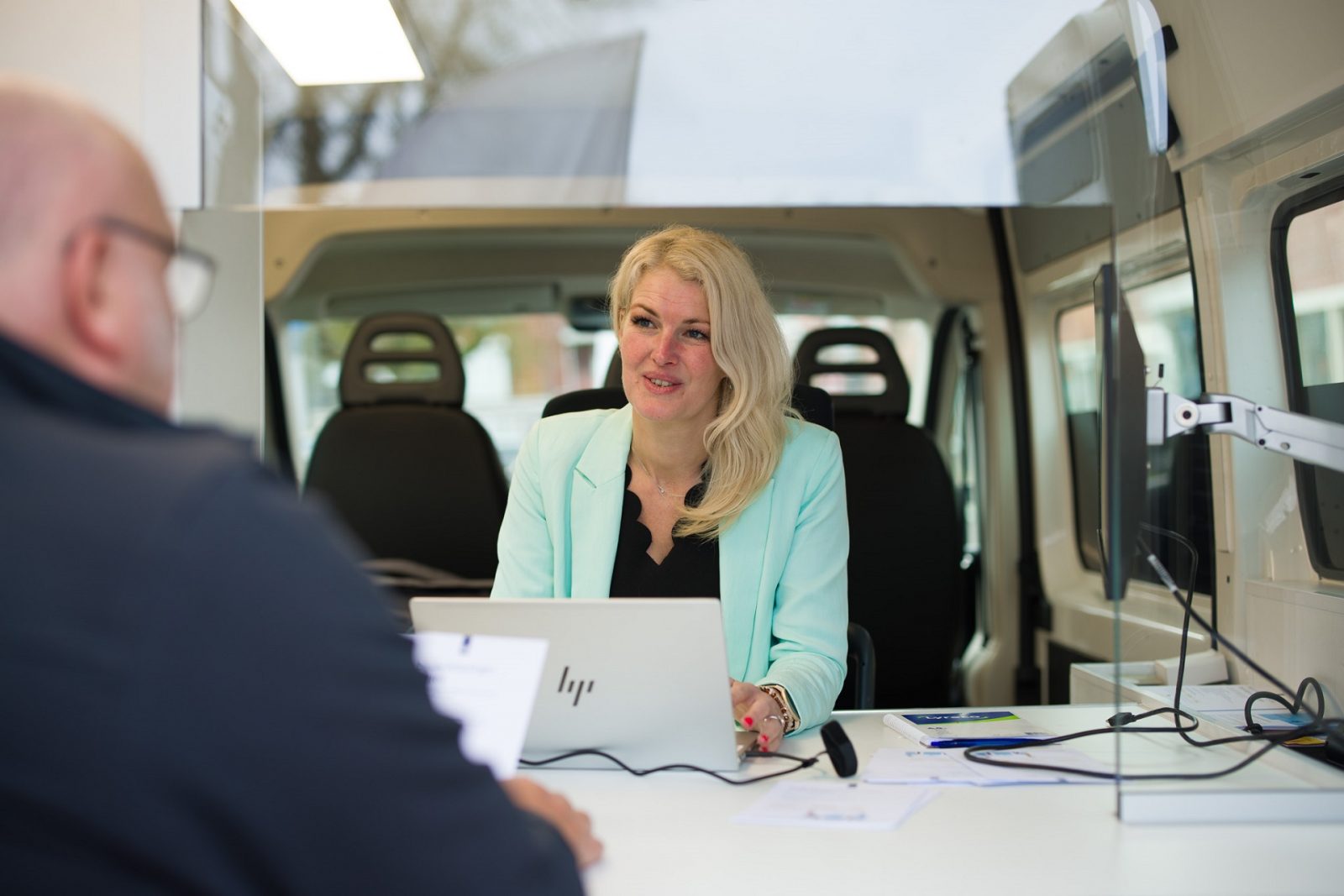 Medewerker dienstverlening Henny van der Wal helpt een bezoeker van de Landelijke Aangiftedag in de Belastingbus. We zien Henny van voren. Ze zit achter haar laptop.